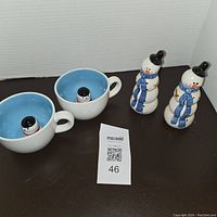 Top-down view of two snowman mugs showing small snowman figures inside, and two matching snowman salt and pepper shakers with black hats and blue scarves.