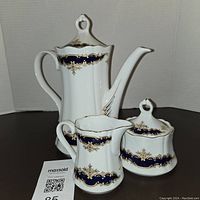 Porcelain teapot, creamer, and sugar bowl on black surface with white background, showing gold swirl and navy blue pattern