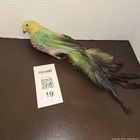 Vintage green parakeet bird tree ornament with yellow head, green body, glitter on the underside, and long natural feather tail with black tips. Shown next to MaxSold lot card