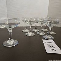 Seven identical smaller vintage mid-century champagne glasses and one slightly larger champagne glass grouped together on a dark surface with a MaxSold tag.