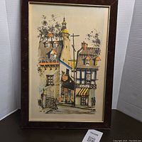 Framed watercolor painting showing streetscape with buildings and church steeple, with visible signature in lower right and label in lower corner.