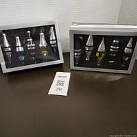 Two original boxes each sealed and containing five glass teardrop-shaped ornaments with striped patterns visible through clear plastic fronts.