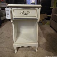 Front view of vintage white nightstand showing drawer with decorative metal handle and open lower shelf.