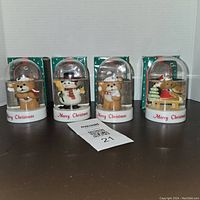 Front view of the four snow globes arranged side by side with Merry Christmas bases and original boxes behind