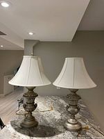 Pair of table lamps with grey bases and white bell-shaped shades on a countertop under ceiling lights.