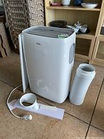 White TCL portable air conditioner with grey control panel, remote, exhaust hose, and window attachments on floor.