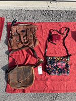 All four bags laid out on red fabric: brown leather satchel (top left), small red wallet with strap (top right), tan leather messenger bag (bottom left), black floral printed Patricia Nash purse (bottom right).