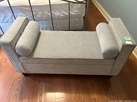 Top-down view of grey upholstered bench with bolster pillows on each side, placed on wooden floor next to bed frame.
