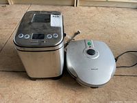 Shows two appliances: the taller rectangular Cuisinart bread maker in stainless steel beside the round Brentwood egg poacher in silver color with indicator lights and knob on top.