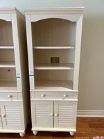 Front view of cream colored wall cabinet showing upper open shelves, middle drawer with two knobs, and double louvered doors below.