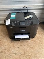 Front view of Canon Maxify MB2720 printer on a floor with paper loaded in the tray and extra ink cartridges on top.