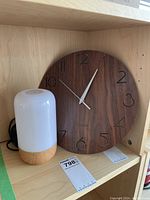 Round wall clock with wood finish and black numbers, white hands, quartz movement next to a cylindrical portable light with wooden base and white frosted glass top.