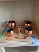 Photo showing copper drink set with two hammered mugs with handles and straws, two round cups, one double-sided jigger, and two copper stir sticks on a wooden shelf.