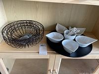 Photo of black tiered wire basket and black lazy Susan with white ceramic bowls and spoons on wooden shelf
