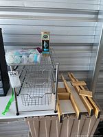 Full view of chrome under-sink storage rack with three wire basket drawers, boxed argan oil bottle, clear bag containing travel containers, and black makeup bag all placed on or beside the unit.