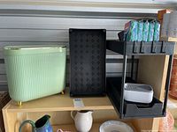 Green retro style waste bin with gold feet, black plastic shelving unit, white ceramic pitcher on shelf below