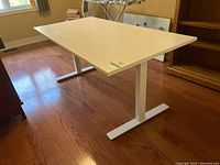 Side view of the large white sewing table showing rectangular top and T-shaped metal legs on hardwood floor.