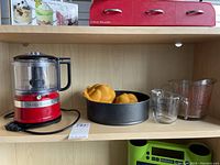 Photo of red KitchenAid food chopper, black cake pan with two orange silicone moulds inside, and two glass measuring cups on a wood shelf.