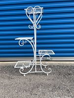 Front view of white metal plant stand showing four shelves and decorative scroll details.
