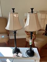 Pair of black metal base table lamps with beige fabric shades shown standing on floor next to upholstered chairs. Lamps have black finials and cords visible, tags attached to cords.