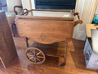 Front and side view of the vintage wooden tea wagon showing decorative carved wood design, large spoked wheels, and handle.