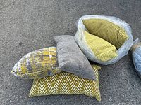 Stack of five throw pillows in grey and mustard yellow tones stacked on pavement, showing various textures and patterns including chevron, geometric lines, and abstract design.