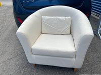 Front and angled front view of a white fabric tub chair with a small matching throw pillow, positioned on asphalt in front of a blue Hyundai Elantra.