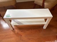 White narrow shelving unit with a smooth finish, seen from above showing the top surface and lower shelf, placed on wooden flooring between two chairs.