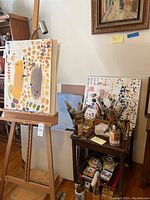 Wooden easel with adjustable supports holding an abstract painted canvas, next to side table with brushes and paint sets