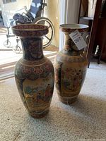Two tall Asian style vases displayed on the floor near a wheelchair, showcasing their full height and decorative painted designs with people and floral motifs.