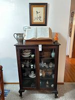 Wooden display cabinet with glass doors closed showing decorative items inside and objects on top including an open book, metal pitcher, and ceramic bird figure