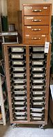 Wooden cabinet with four labeled drawers and metal trays below, showing various hardware items organized in trays.