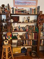 Full view of wooden shelving unit filled with books, collectibles, masks, pottery and decorative items. Includes a wooden stool with a ceramic jug in front.