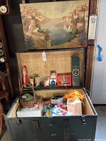 Vintage metal trunk open with assorted holiday decorations inside and framed painting on top