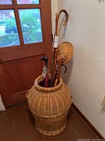 Large wicker basket at wooden door corner holding wooden walking sticks and umbrella, showing overall lot composition.