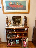 Shelving unit filled with assorted art decor, wooden sculptures, thimble display case and boxed items beneath a framed print of dogs in a boat