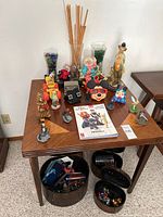 Wide view of wooden card table with clown figurines, Mickey Mouse item, glass vases filled with marbles, and incense sticks. Nested black metal storage containers filled with small collectibles visible below table.