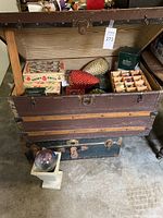 Open steamer trunk lined with striped fabric showing assorted ornaments, stacked on smaller trunk with glass orb on pedestal in front