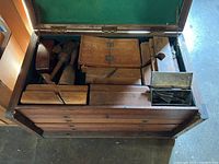 Top view of open wooden tool chest showing green felt-lined interior and compartments filled with wooden molding planes and metal plane irons