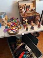 Large assortment of vintage toys on table: Lincoln Logs can, Tinker Toys parts scattered, Lego bricks in round tin, various wooden figurines on table and inside trunk.