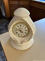 Porcelain acorn-shaped clock with gold-trimmed round dial showing Roman numerals and Belleek branding in the center, photographed standing on a counter in natural light.