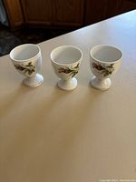 Three white ceramic egg cups with unique floral designs. Each cup has a pedestal base and appears in good condition.