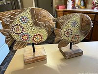 Two wooden fish sculptures with mosaic tile patterns on a kitchen counter, showing side views and bases.