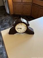 Front angled view of Irish bog oak clock on a counter with kitchen cabinets in the background. The clock has a circular white face with Roman numerals and decorative carved wood body.