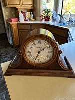 Wooden Howard Miller mantle clock with rounded wooden casing placed on white surface, kitchen background visible.