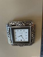 Frontal view of the silver filigree travel clock showing the square shape, detailed silver frame pattern, and clock face with numbers and hands visible