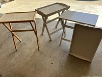 Three TV trays in gray and white colors shown standing on a concrete floor with folding legs extended.