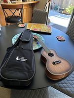 Photo of Cordoba 15CM-E ukulele lying on black dining table next to Ted Brown Music branded black soft case. Table and chairs visible in background.