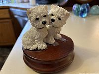 Two West Highland White Terrier figurines modeled closely on a dark wooden circular base. The detailed cream-colored dogs have textured fur and black facial features.