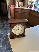 Wood grain mantel clock with cream face featuring Roman numerals and black trim, placed on white countertop with kitchen background.